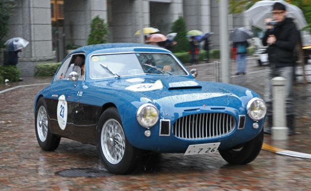 Fiat 8V bleue engagée dans une course historique sous la pluie, roues à rayons et numéro de course visible