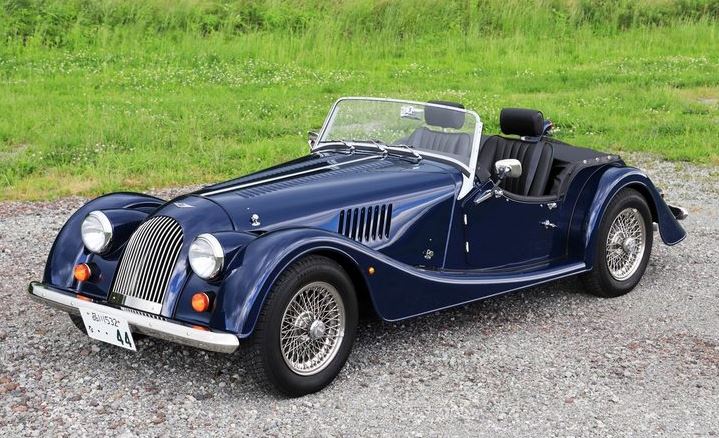 Morgan Plus 4 bleue stationnée sur gravier au bord d’un paysage verdoyant