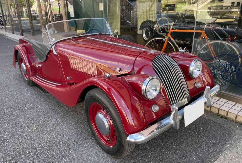 Morgan Plus 4 rouge des années 1950 stationnée devant des vitrines urbaines