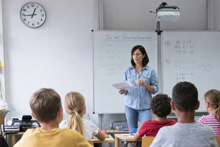 Salle de classe avec une horloge analogique au mur, une enseignante au tableau et des élèves assis