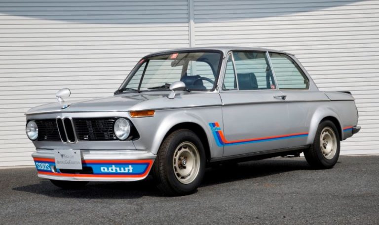 BMW 2002 Turbo grise vue de face trois quarts avec bandes Motorsport sur route devant fond neutre