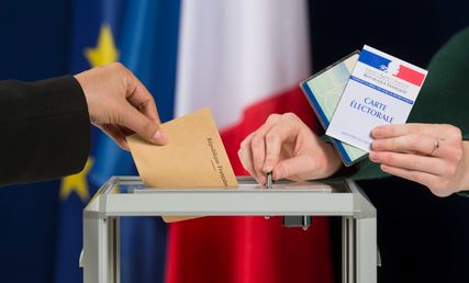 Main d’un électeur glissant un bulletin dans l’urne lors d’une élection municipale en France, avec carte électorale et drapeaux français et européen en arrière-plan.