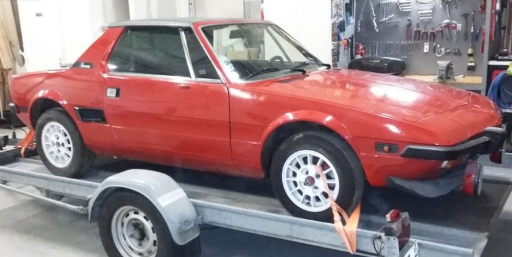 Fiat X1/9 rouge de 1978 sur remorque dans un atelier, jantes blanches et carrosserie propre