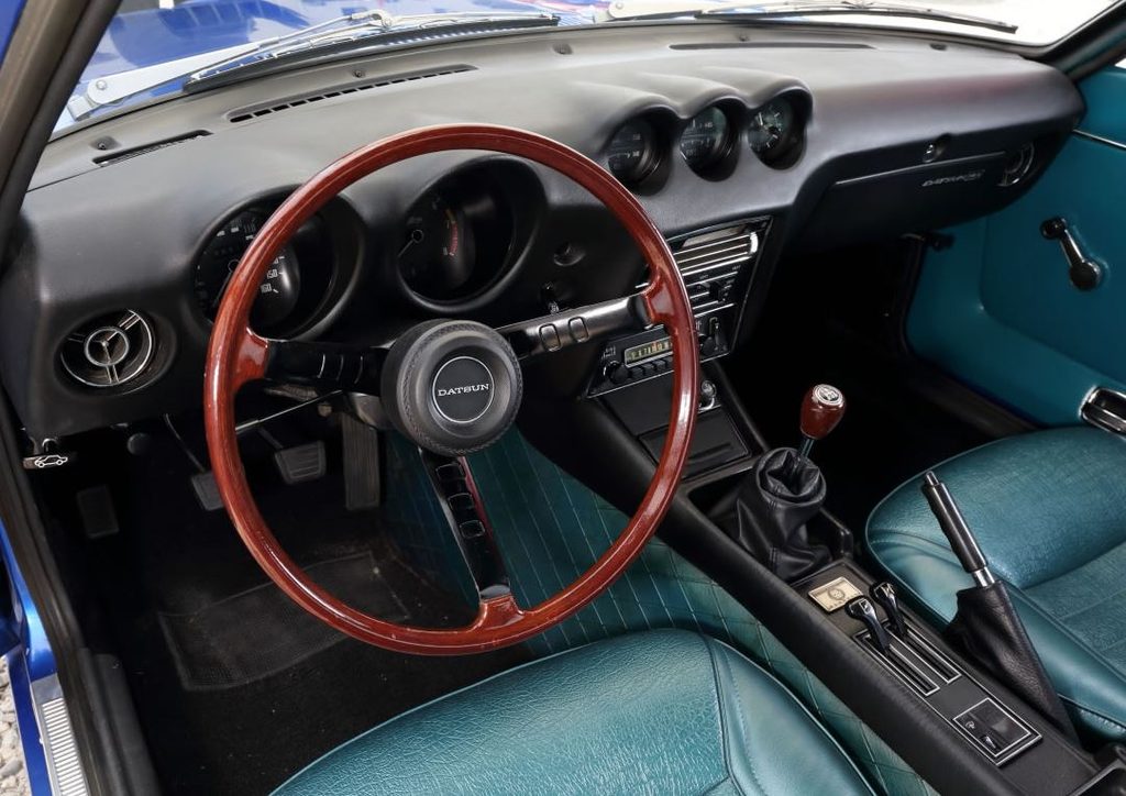 Intérieur de Datsun 240Z avec volant en bois, tableau de bord à trois cadrans centraux, boîte manuelle et sièges turquoise.