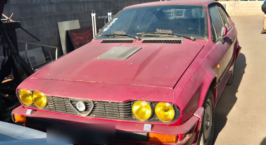Alfa Romeo GTV6 1984 rouge fatiguée avec phares jaunes en sortie de grange