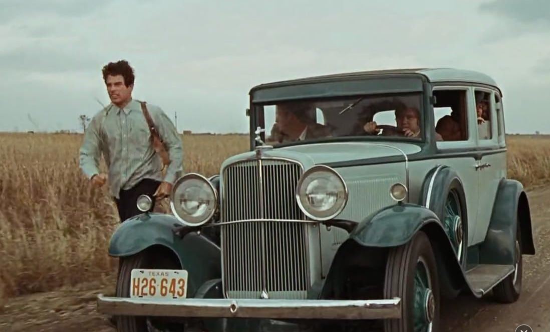 Clyde à côté d’une Ford V8 dans un champ aux États-Unis années 1930