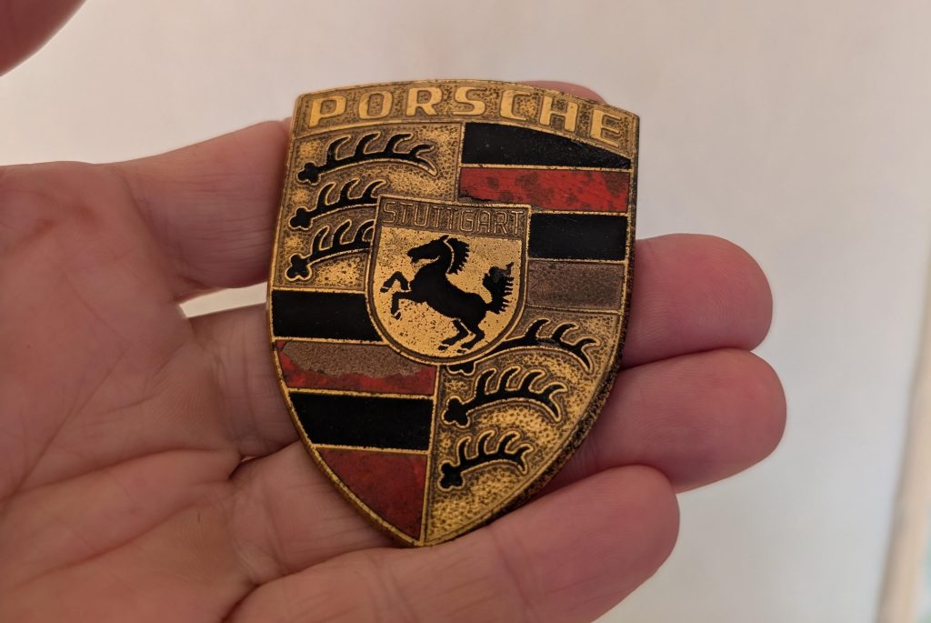 Ancien logo Porsche d’époque tenu en main avec émail usé et rouge vif des premières versions