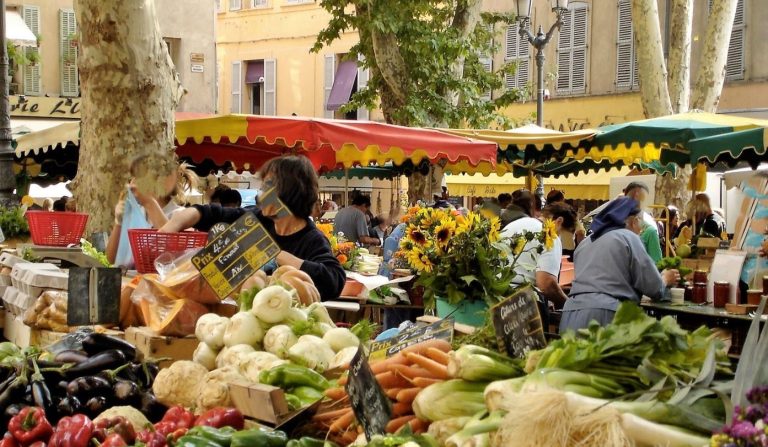 Marché de plein air animé avec étals de légumes frais, commerçants et habitants échangeant sur une place de village ombragée.