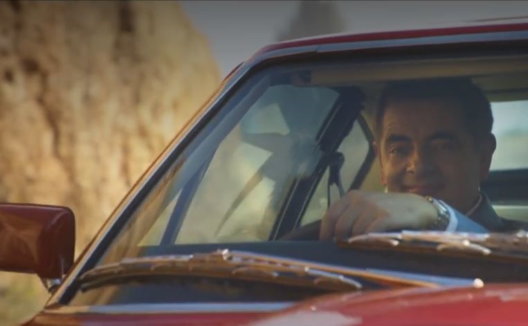 Rowan Atkinson au volant d'une Aston Martin DBS rouge dans une scène de film