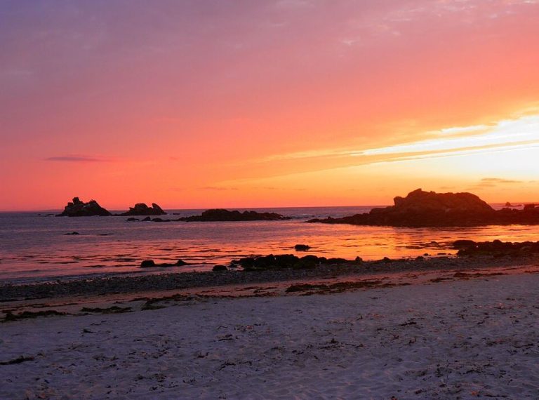 Coucher de soleil sur une plage bordée de rochers, où les couleurs chaudes du ciel se reflètent sur la mer, illustrant la beauté d'un spectacle naturel quotidien.
