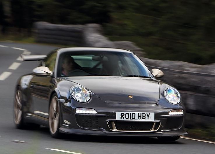 Porsche 911 GT3 RS génération 997 gris foncé photographiée de trois quarts avant sur une route sinueuse.
