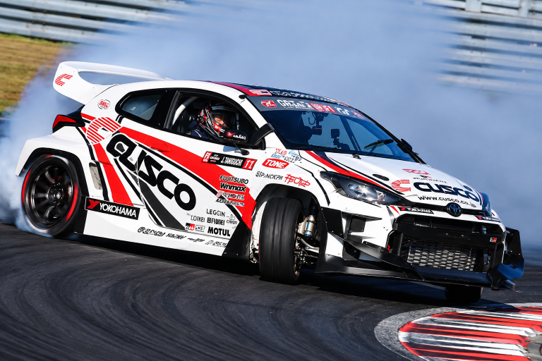 Voiture de compétition blanche aux couleurs Cusco en plein drift sur circuit, avec un contre-braquage extrême et de la fumée.
