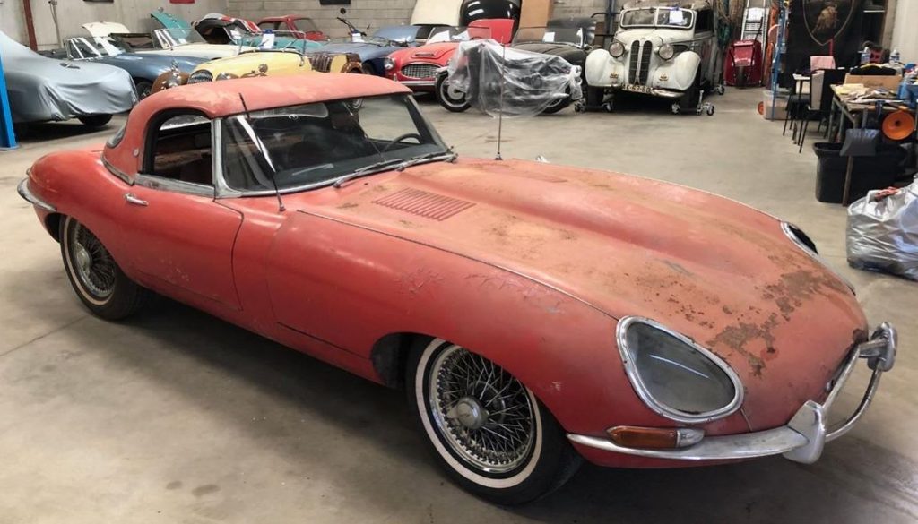 Jaguar Type E cabriolet 1961 à plancher plat, rouge et poussiéreuse, retrouvée dans un garage parmi d'autres voitures anciennes.