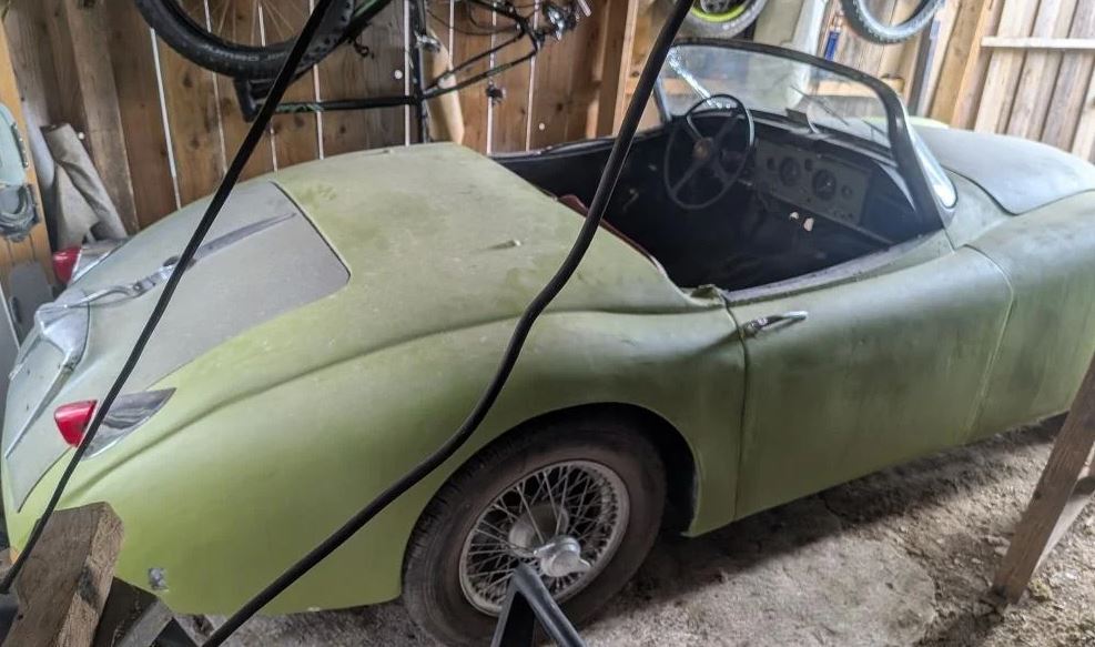 Jaguar XK 150 Roadster de 1959 verte, recouverte de poussière et entreposée dans une grange avec sa capote ouverte.