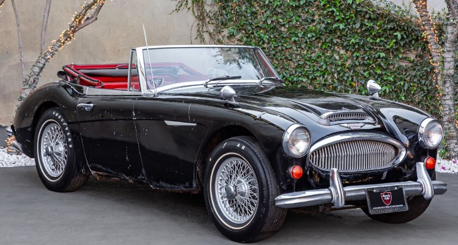 Austin-Healey 3000 BJ8 Mk III de 1964 noire, capote repliée et intérieur rouge, avec traces d'usure sur la carrosserie.