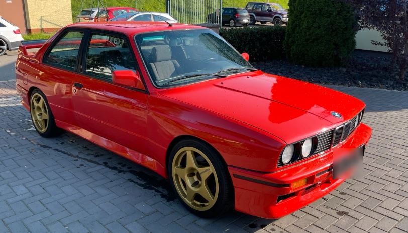 BMW M3 E30 de 1986 rouge, avec jantes dorées et aileron arrière, photographiée de trois quarts avant.