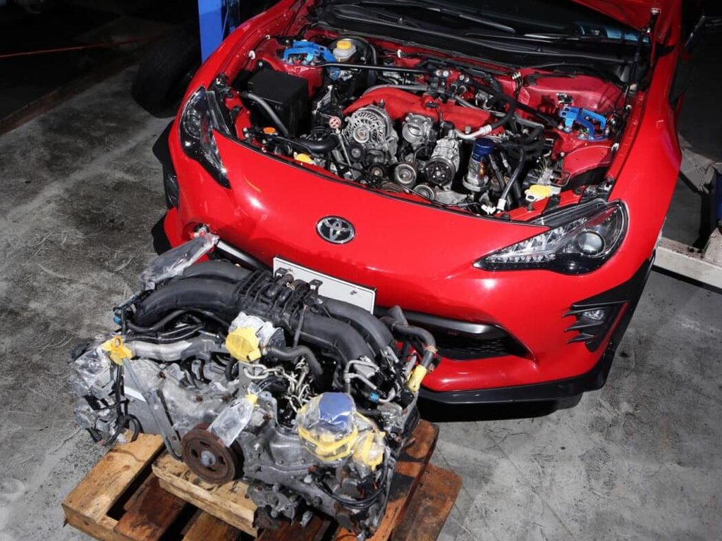 Toyota GT86 rouge dans un atelier, capot ouvert, avec un moteur Boxer quatre cylindres démonté et posé sur une palette.